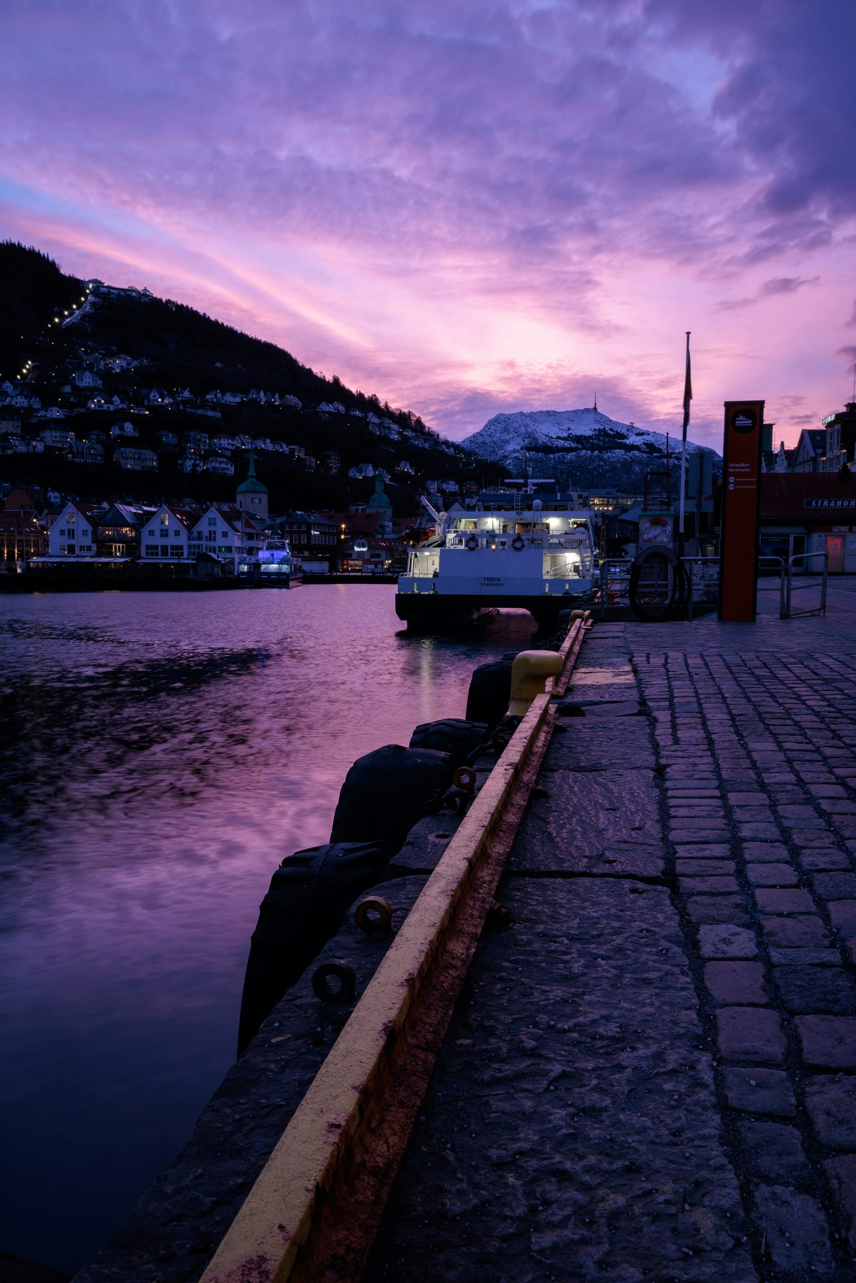 Boats moored in Bergen harbour at dusk with waterfront buildings and lights reflected in the water