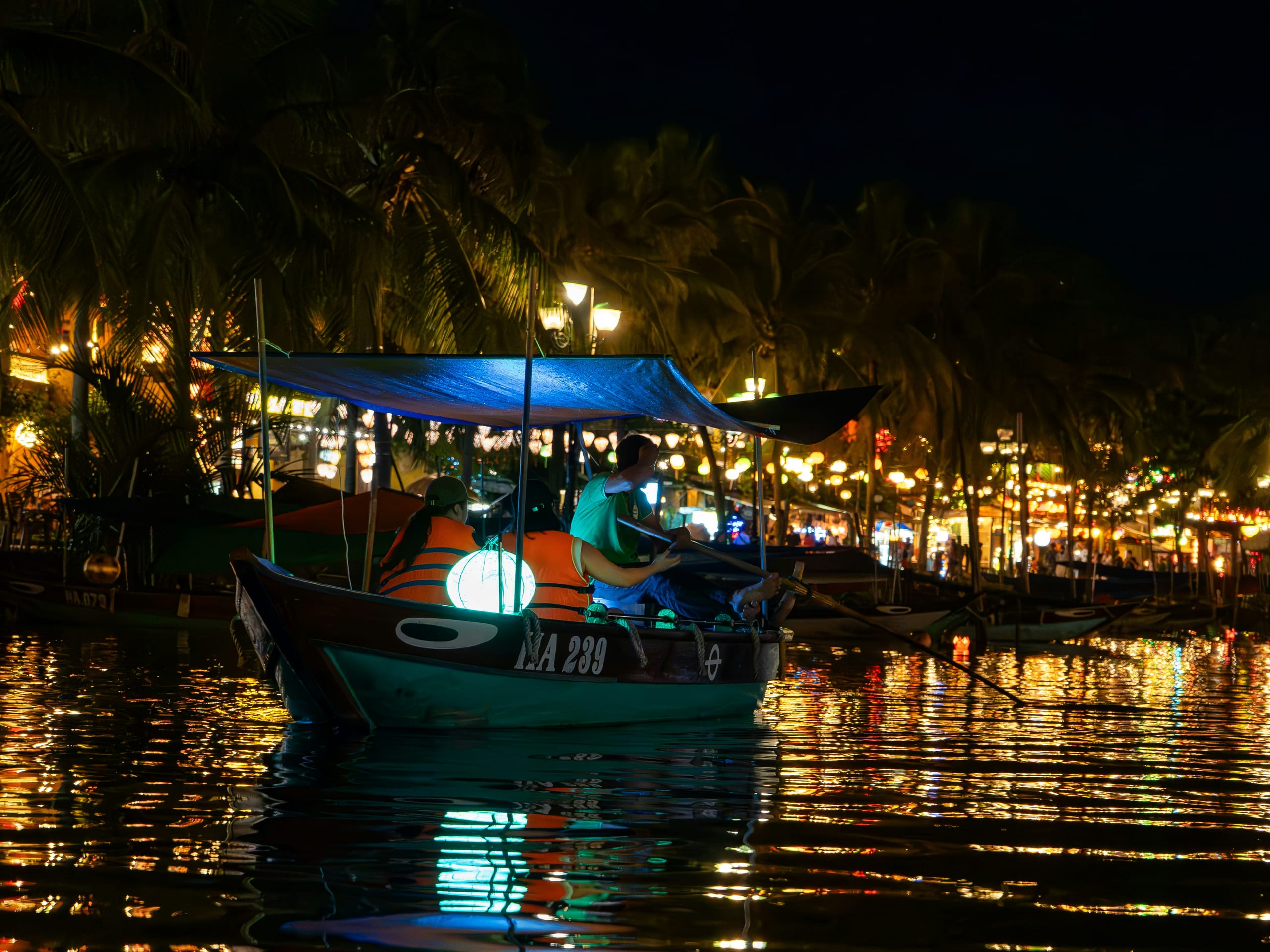 A wooden sampan lit with colored lanterns drifting on the Thu Bon River at night in Hoi An