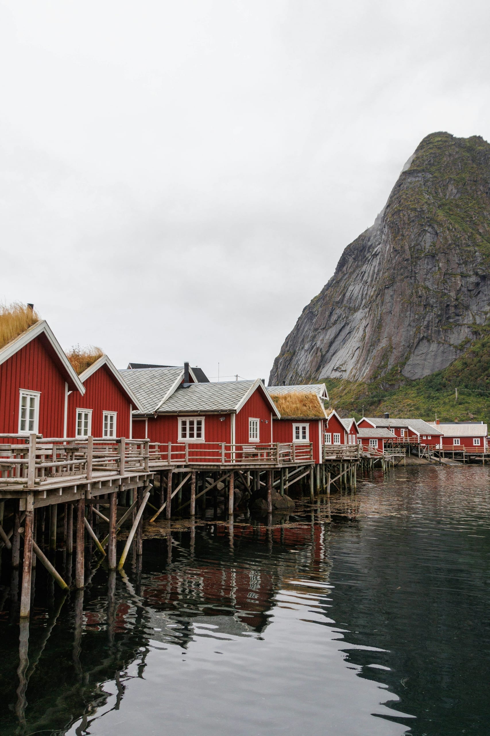 Red fishing cabins on stilts at the water's edge in Reine, with sheer granite peaks rising behind the fjord